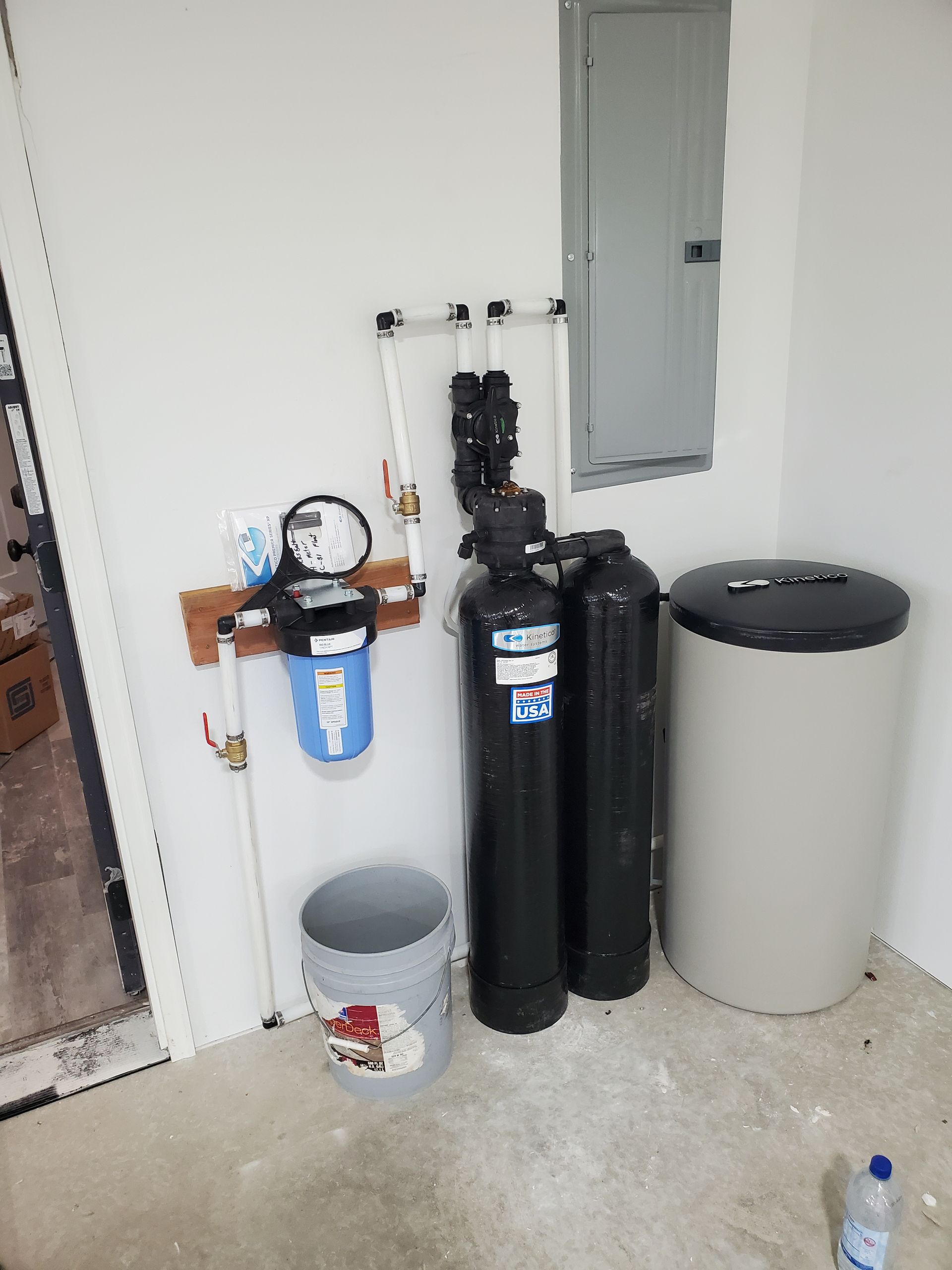 Water filtration system and water softener in a utility room. White and black tanks, and a grey bucket.
