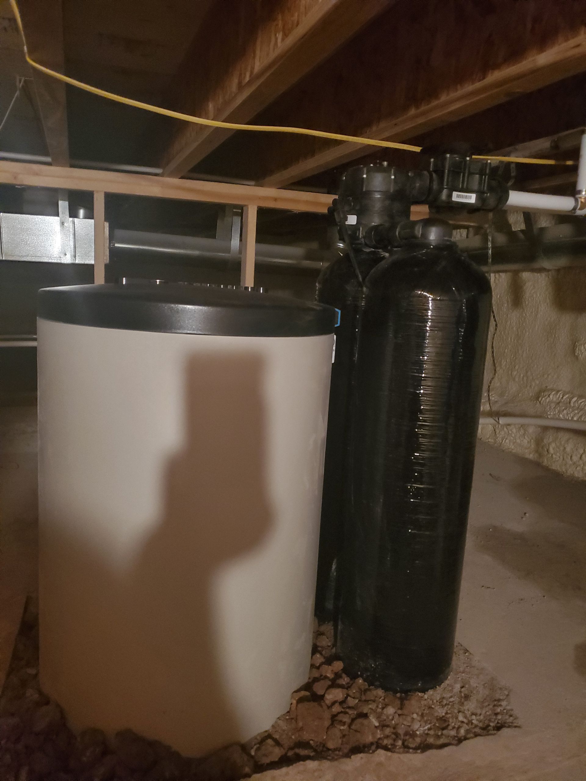 A water softener system in a basement with a white tank and a black tank on a gravel base.