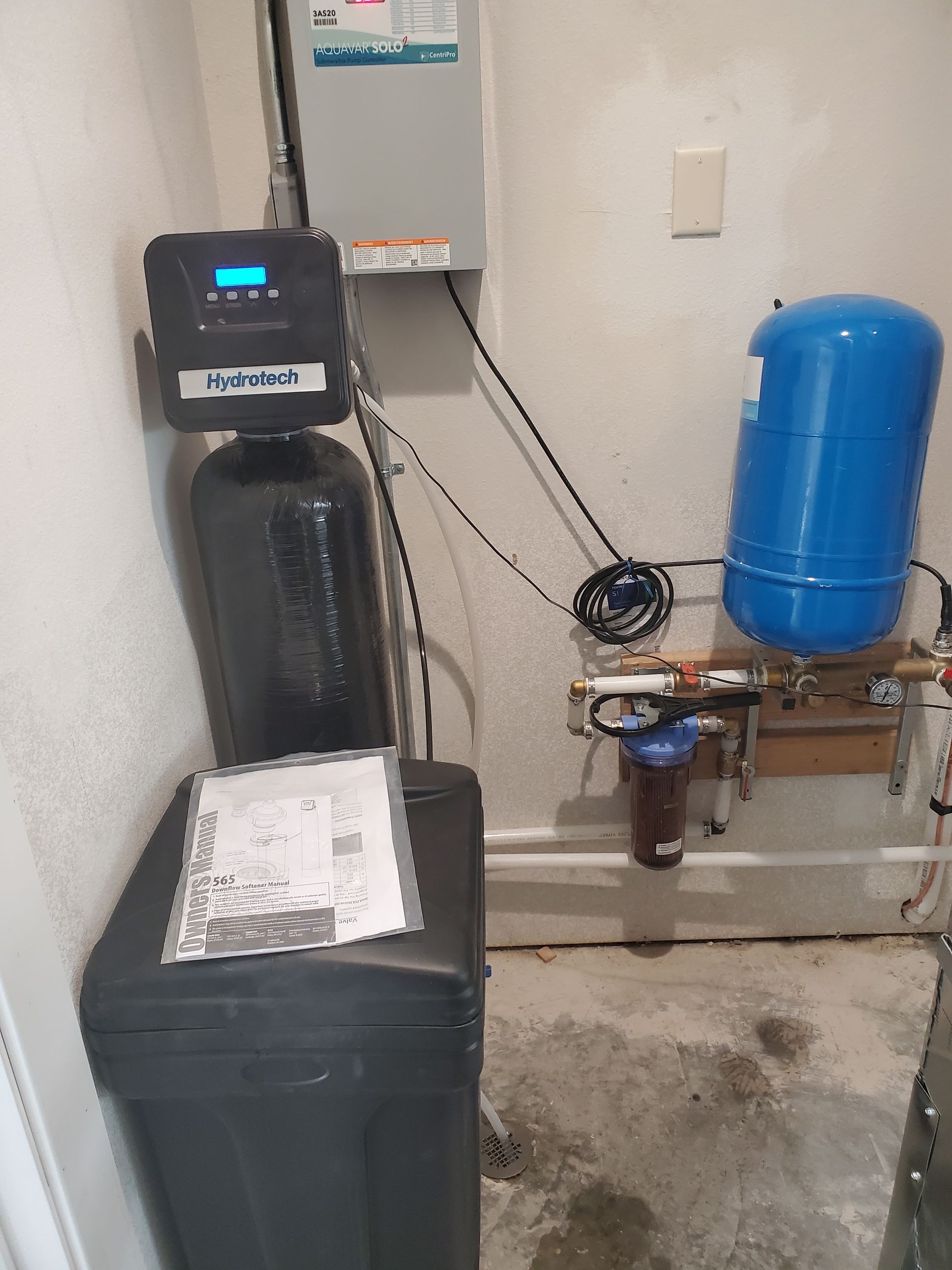 Water softener system in a utility room. Black and blue tanks, white pipes, and a control panel are visible.