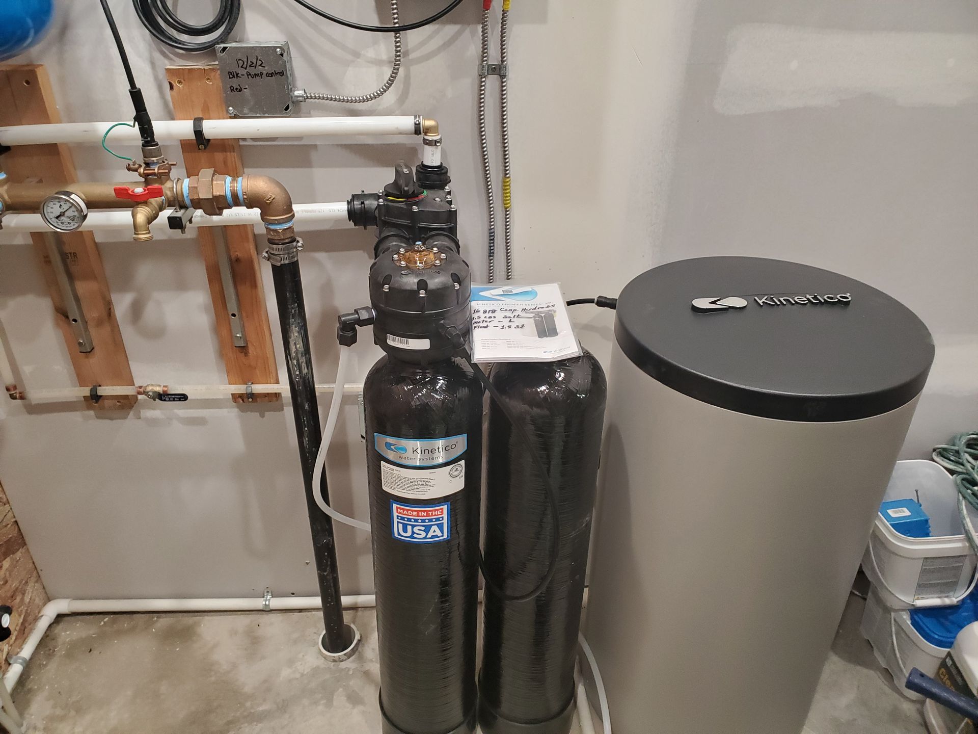 Water softener system in a utility room. Black tanks, gray tank, and plumbing against a white wall.