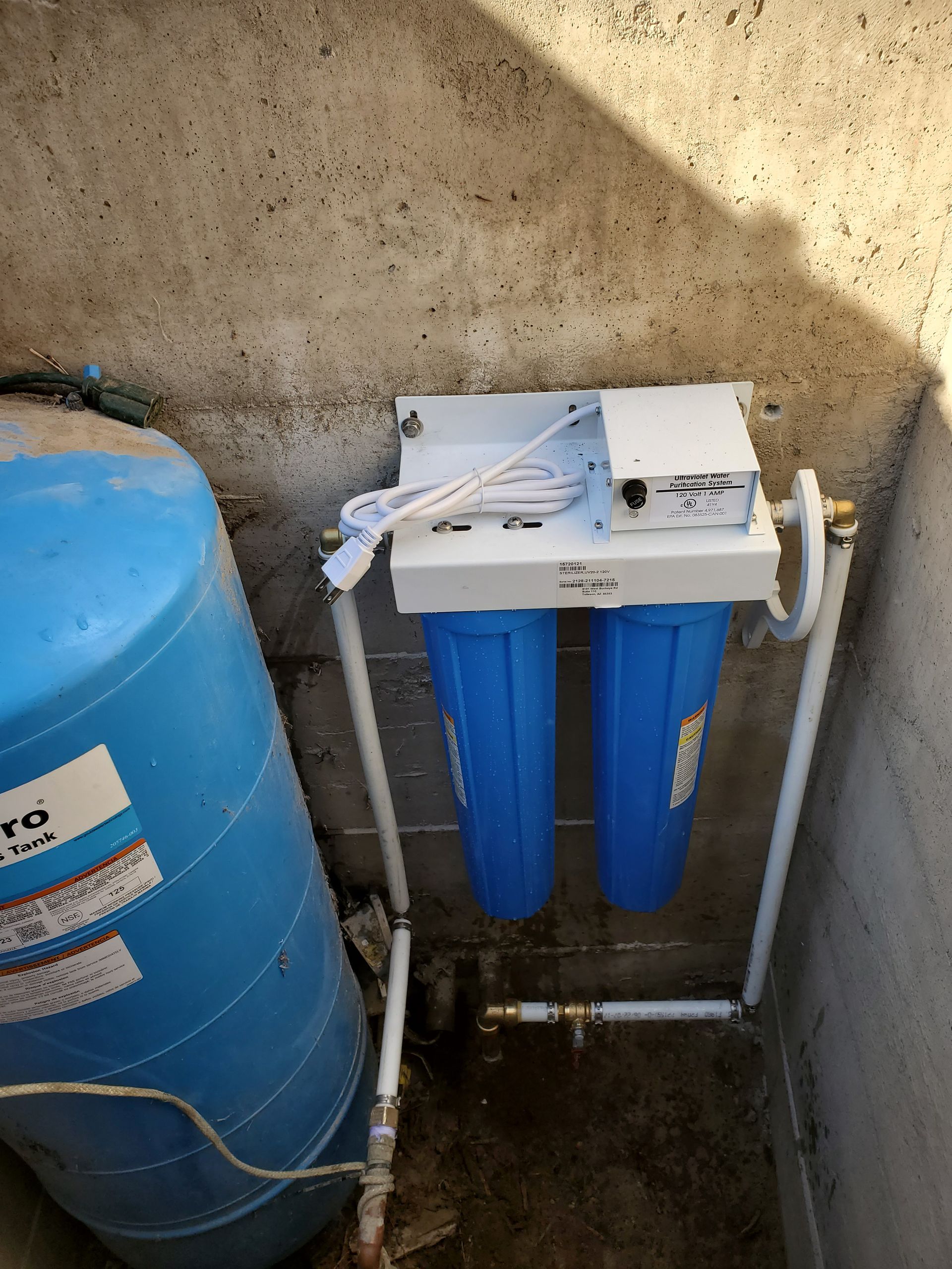 Blue water tank and filter system mounted on a wall. Pipes connect the tank to the filter and onward.