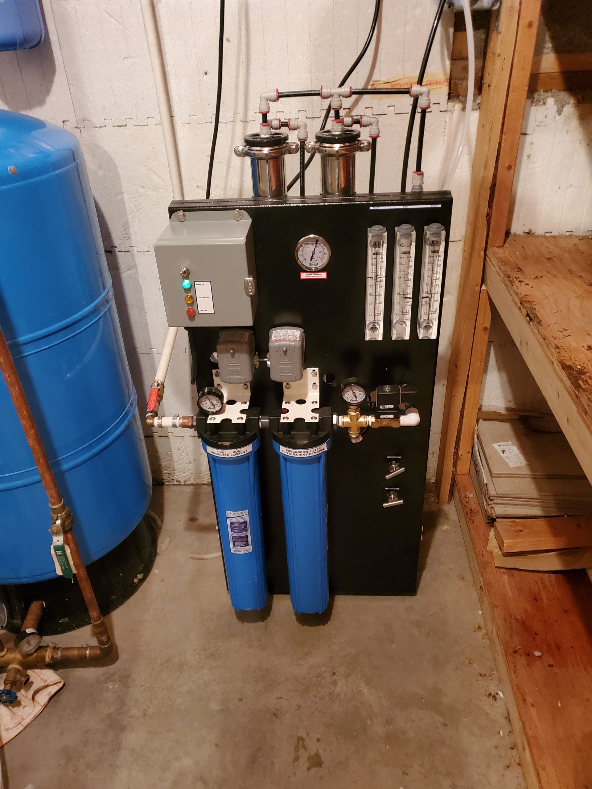 Water filtration system with blue tanks, gauges, and electrical components, against a basement wall.
