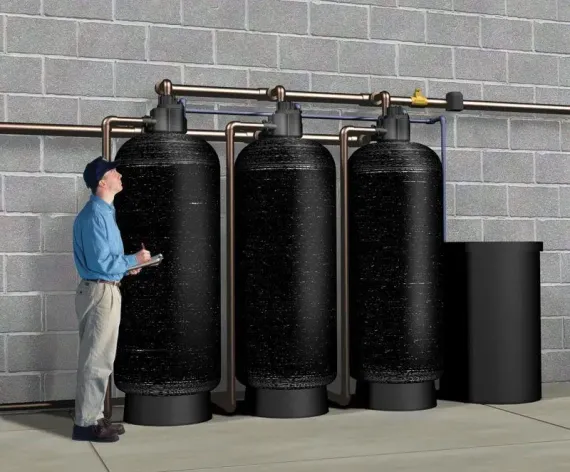 Man inspecting water filtration system; three tall black tanks, pipes, and a black bin against brick wall.