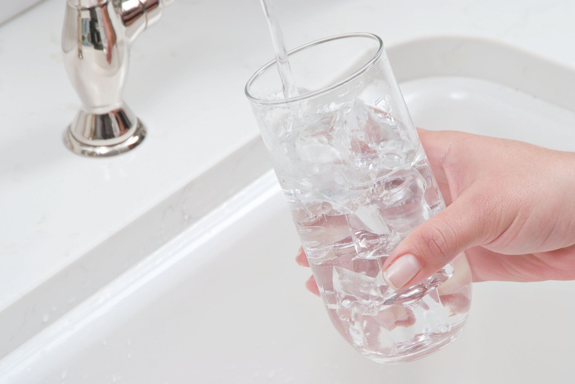 A hand fills a glass with water and ice cubes in a white sink, under a silver faucet.