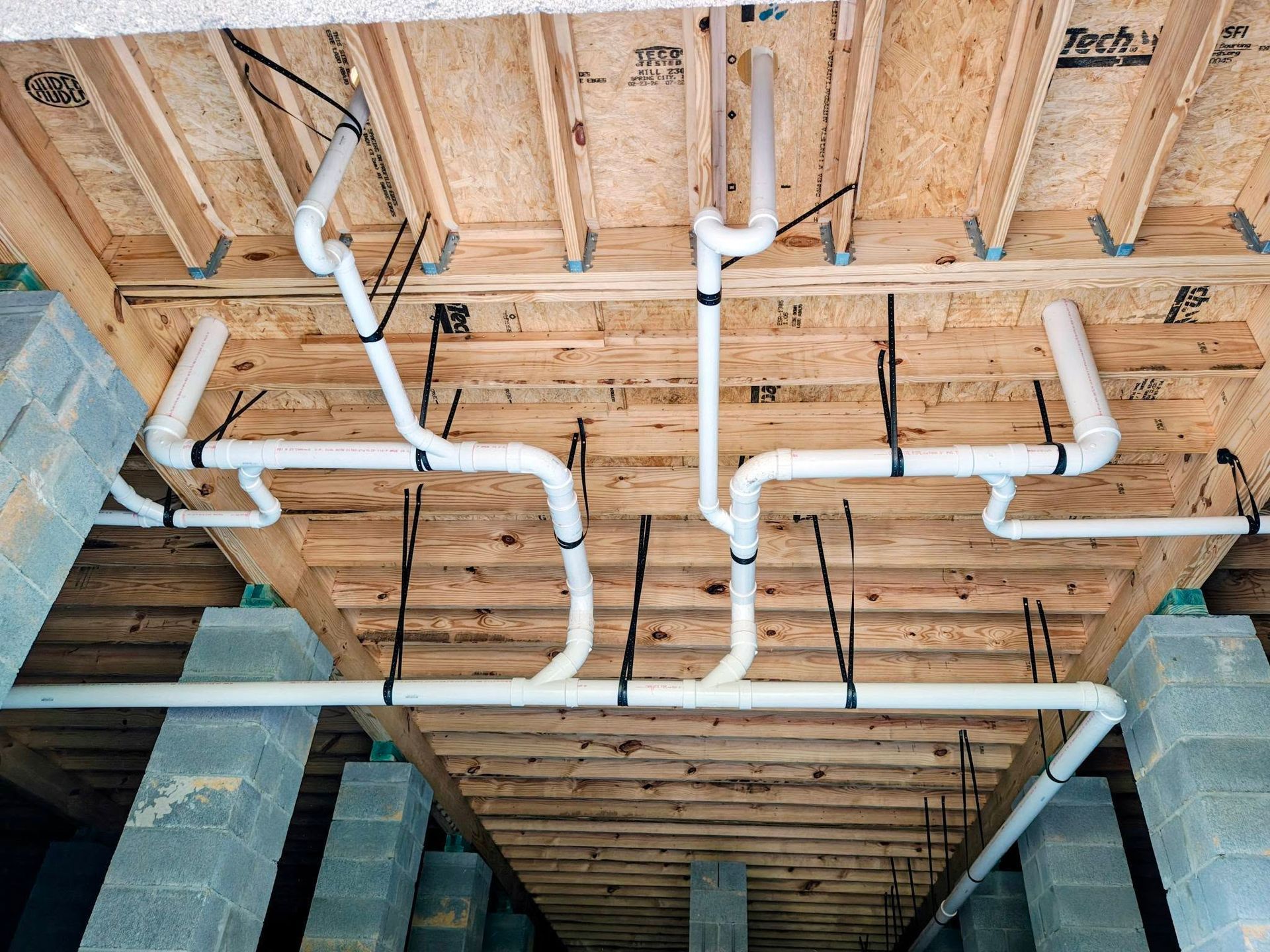 Exposed white PVC pipes and black wiring beneath a wooden floor during construction