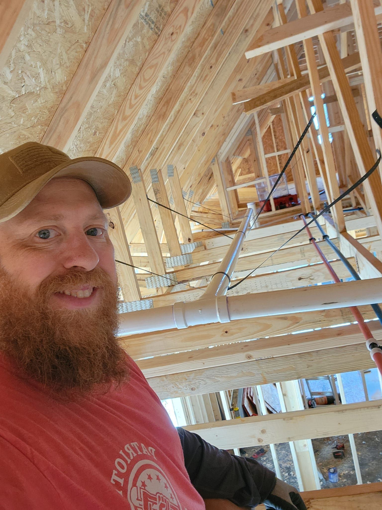 Bearded man in a red shirt and cap inside a wooden house frame under construction