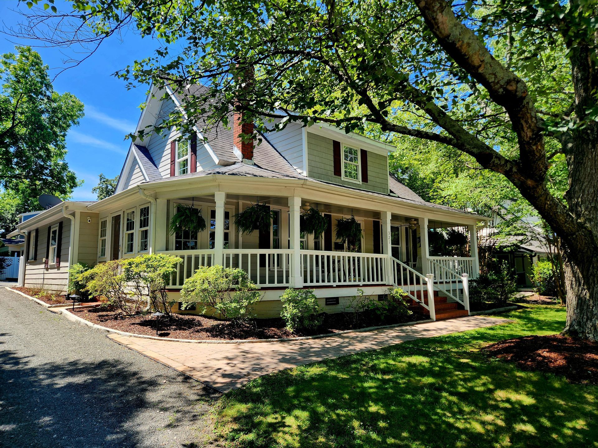 Two-story, light yellow house with wraparound porch, green lawn, and large tree in sunny setting.