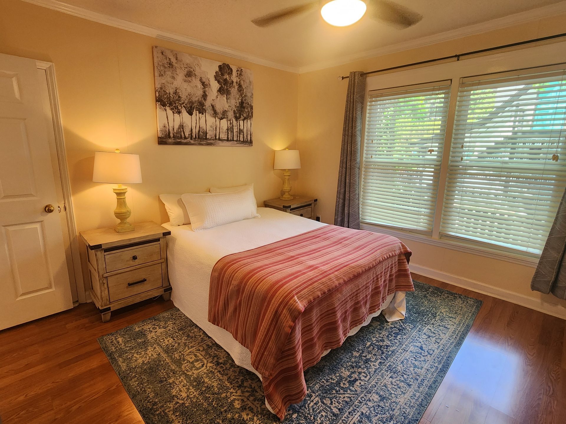 Bedroom with a bed, nightstands, rug, window, and wall art. Warm lighting and wood flooring.