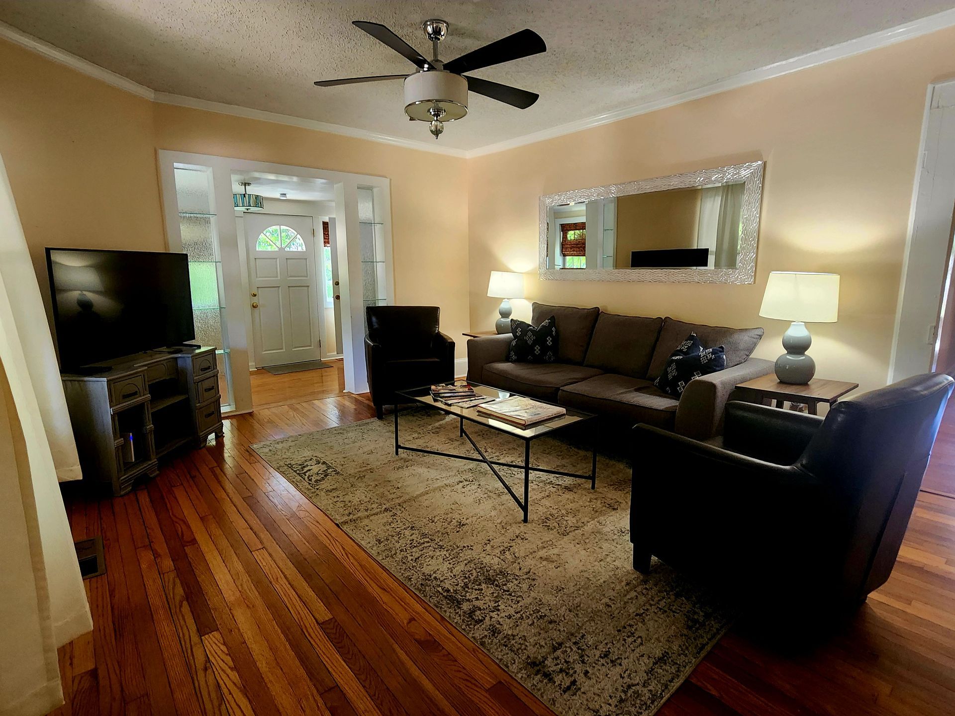 Living room with brown sofa, two armchairs, rug, TV, and ceiling fan. Wooden floor and beige walls.