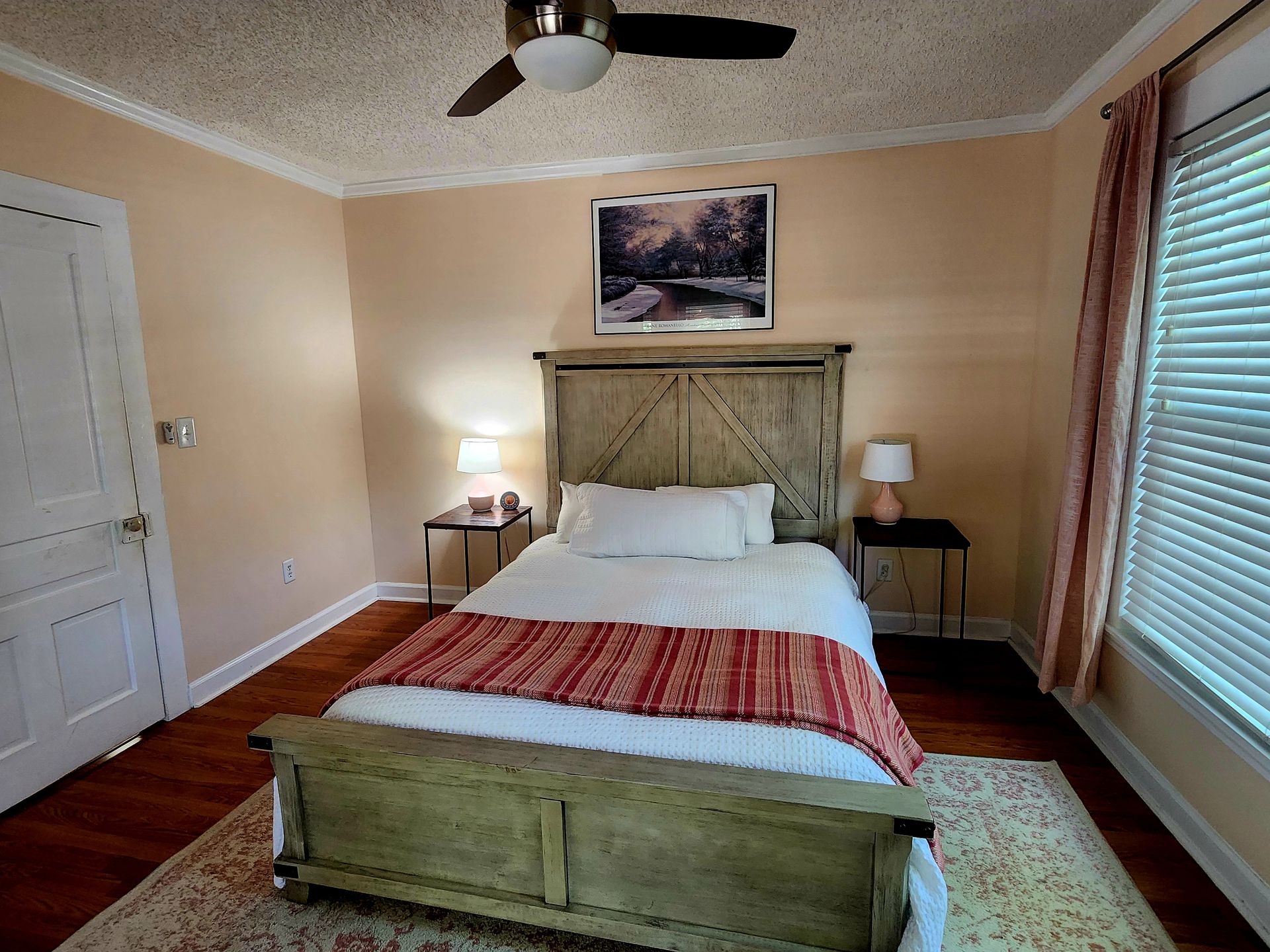 Bedroom with a wooden bed, nightstands, a painting, and a ceiling fan. Light peach walls, wood floor.