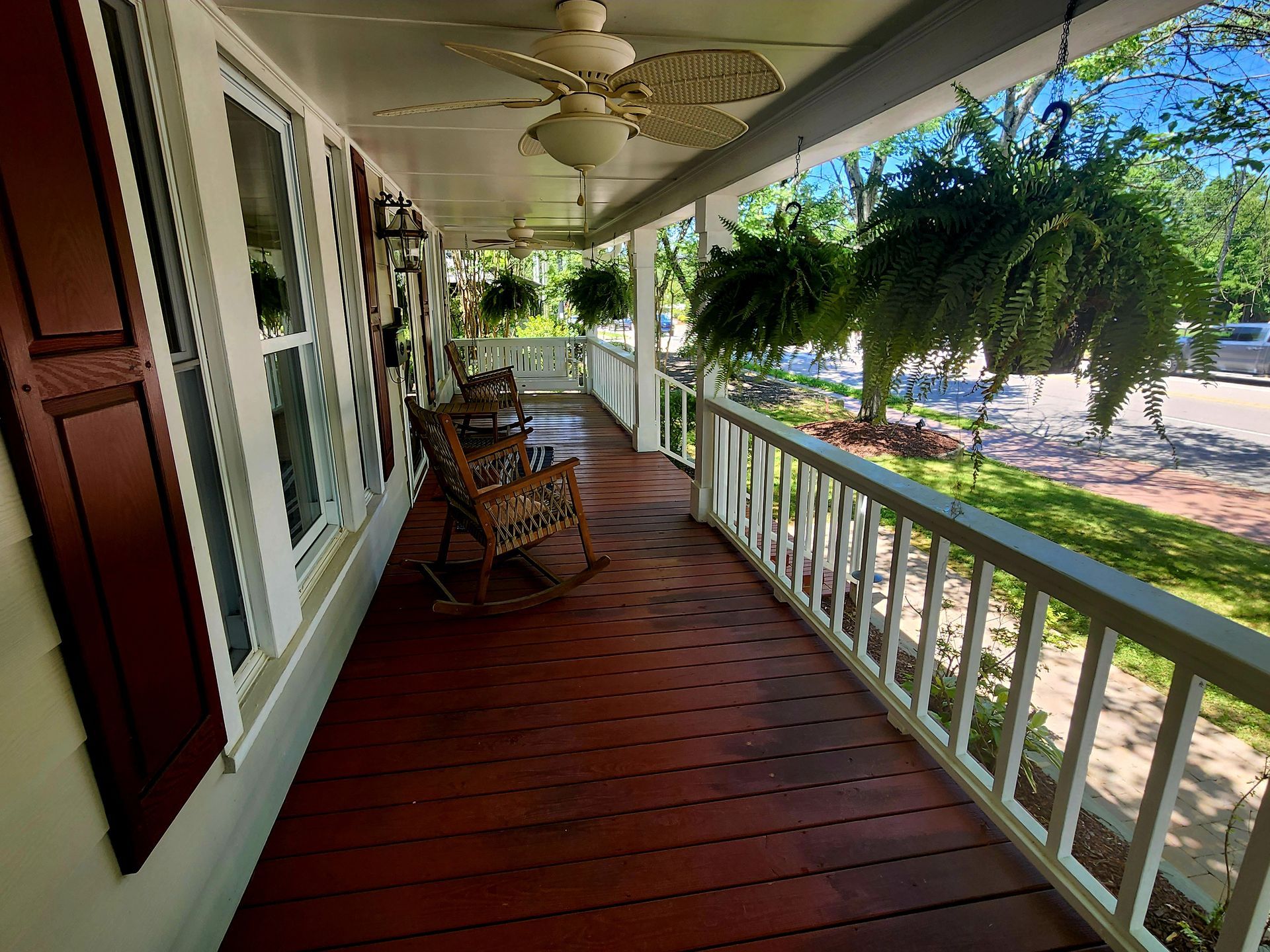 Covered porch with red-stained wooden floor, white railing, and rocking chairs. Ferns hang from the ceiling.