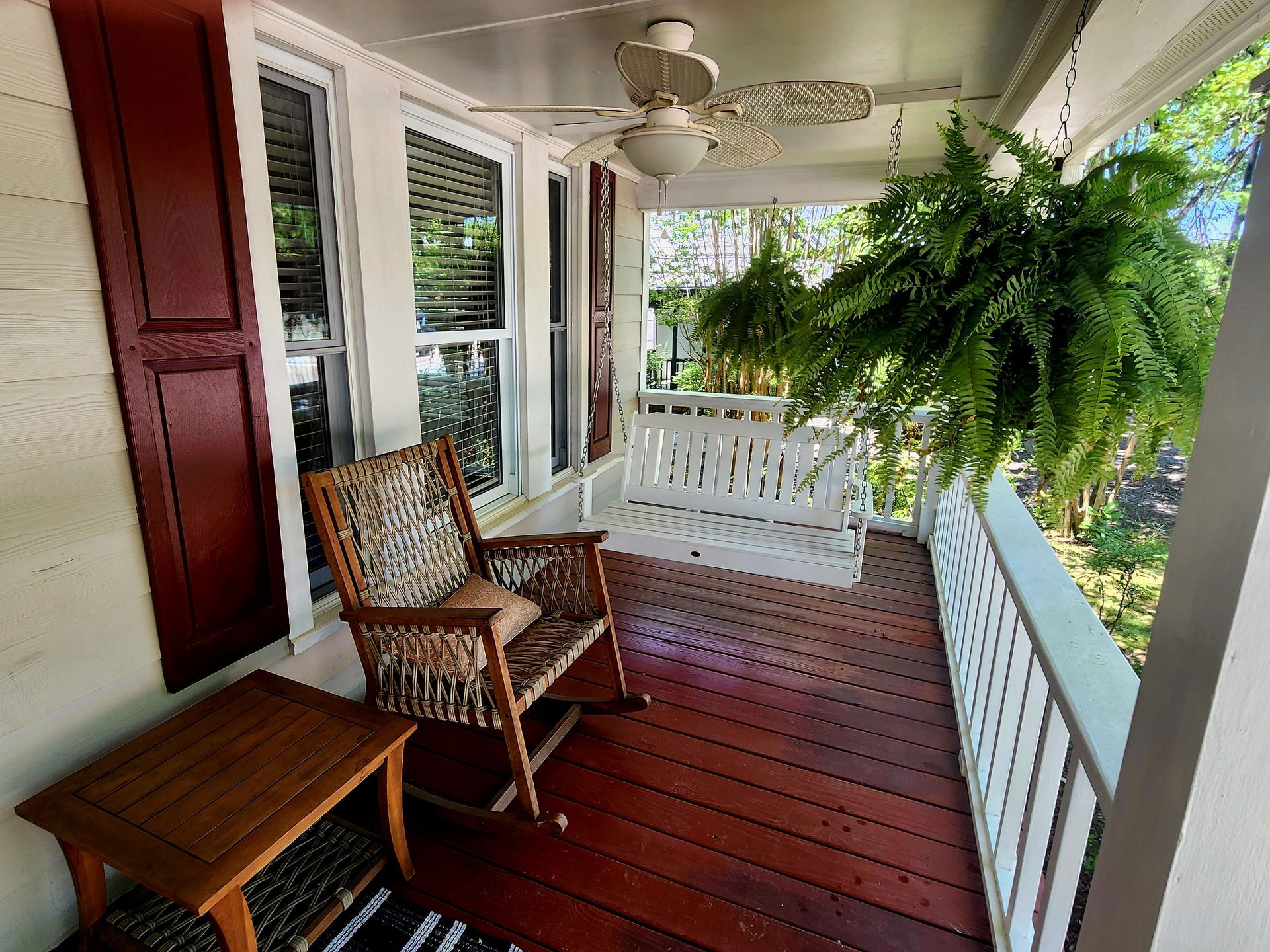 Porch with a rocking chair, swing, and hanging fern. Reddish-brown wood floor. White railings and ceiling fan.