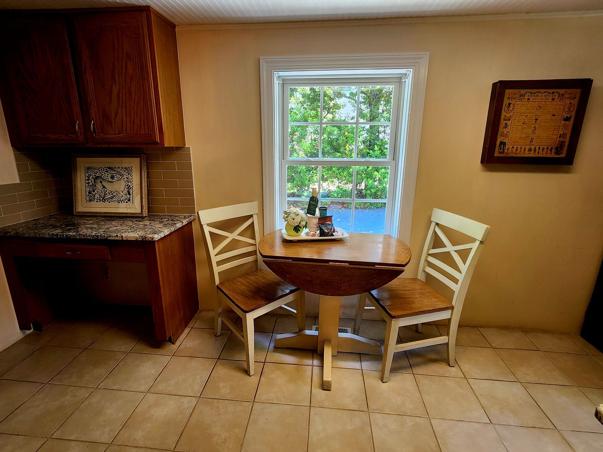 Small kitchen with round table and two chairs near a window, brown cabinets and beige tile.