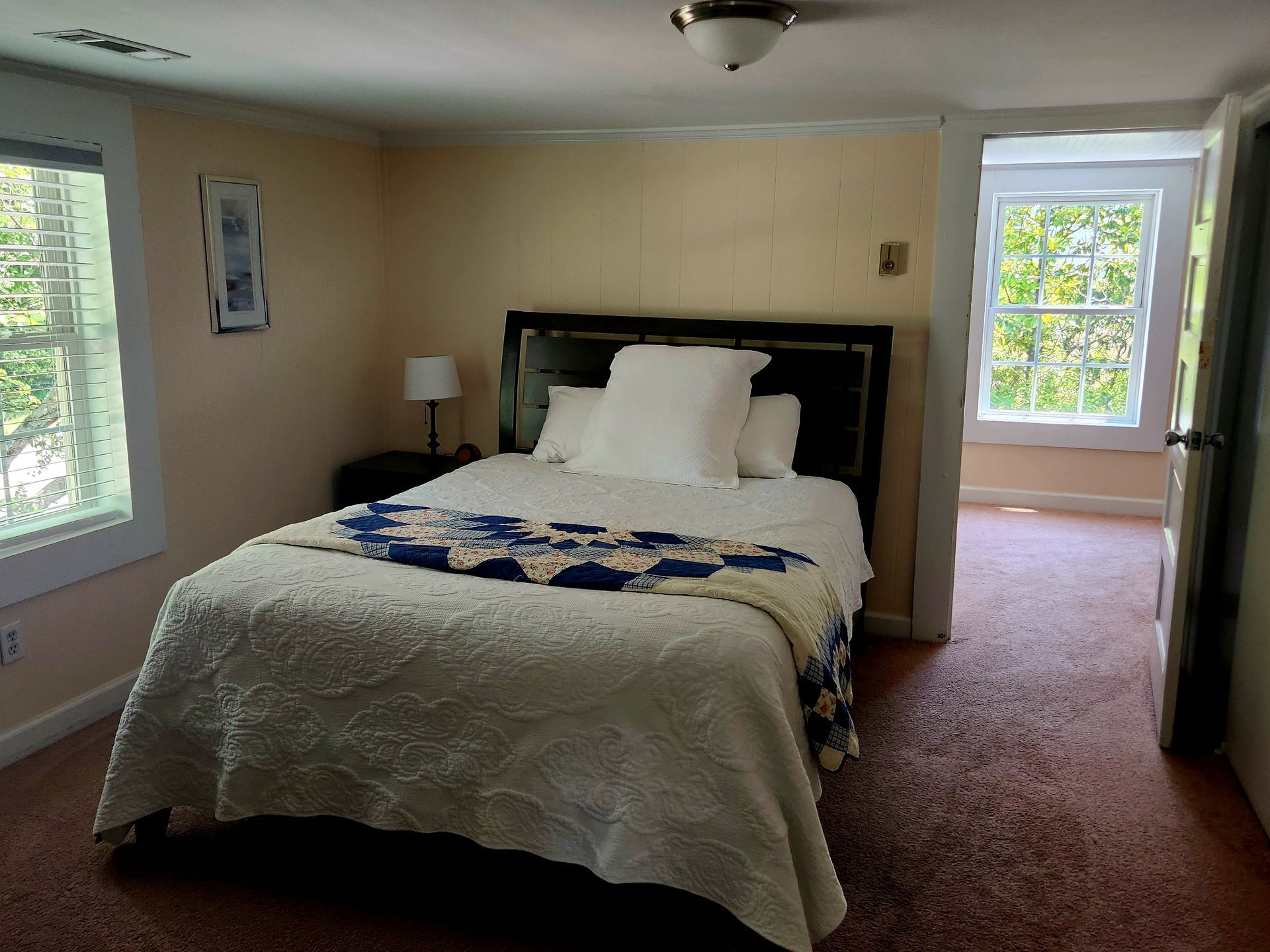 Bedroom with a bed, nightstand, and two windows. Beige walls, brown carpet.
