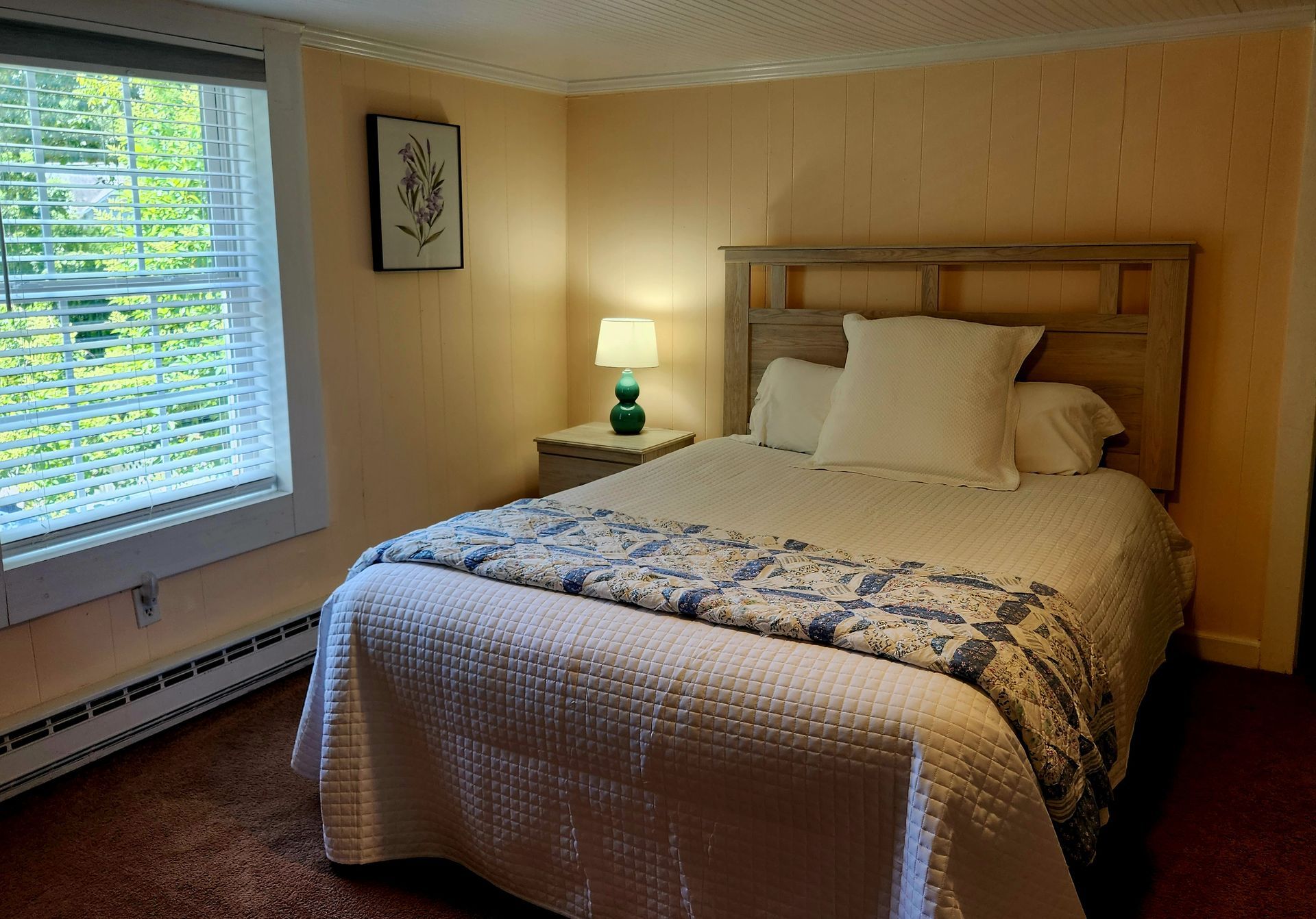 Bedroom with a double bed, nightstand, window with blinds, and artwork. Warm lighting.
