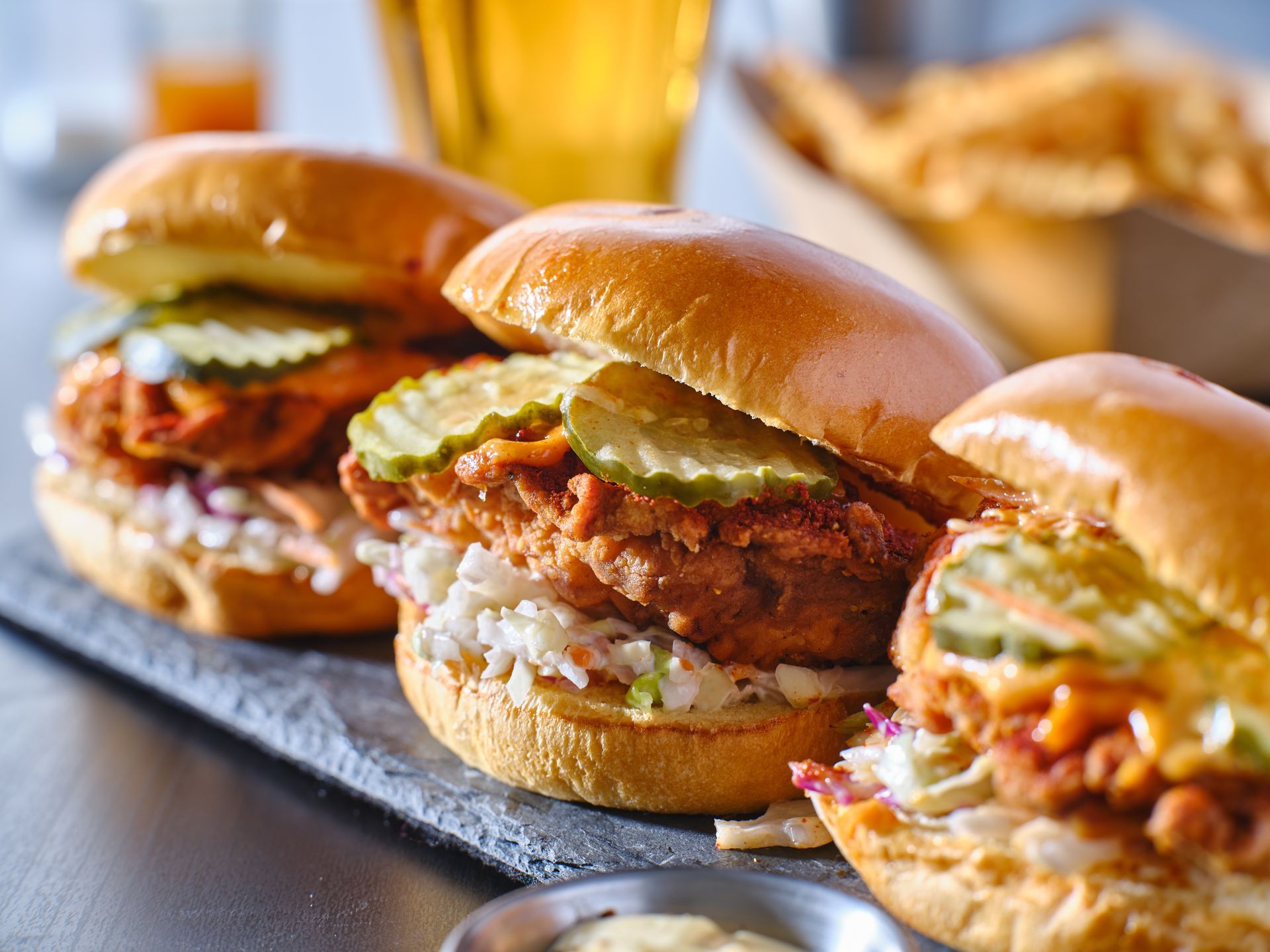 Three fried chicken sliders with pickles, coleslaw, on a slate with fries in the background.