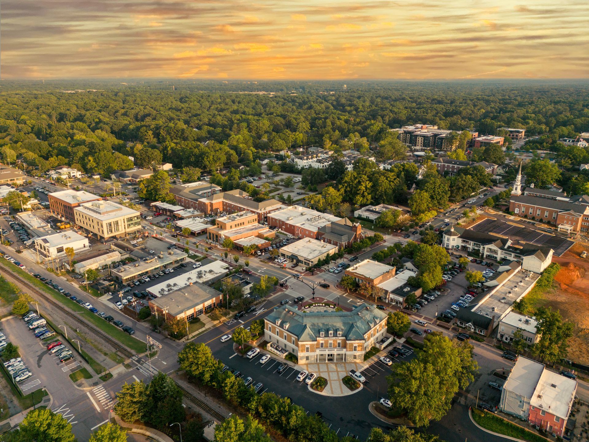 Aerial view of a town square with brick buildings and lush trees under a sunset sky.