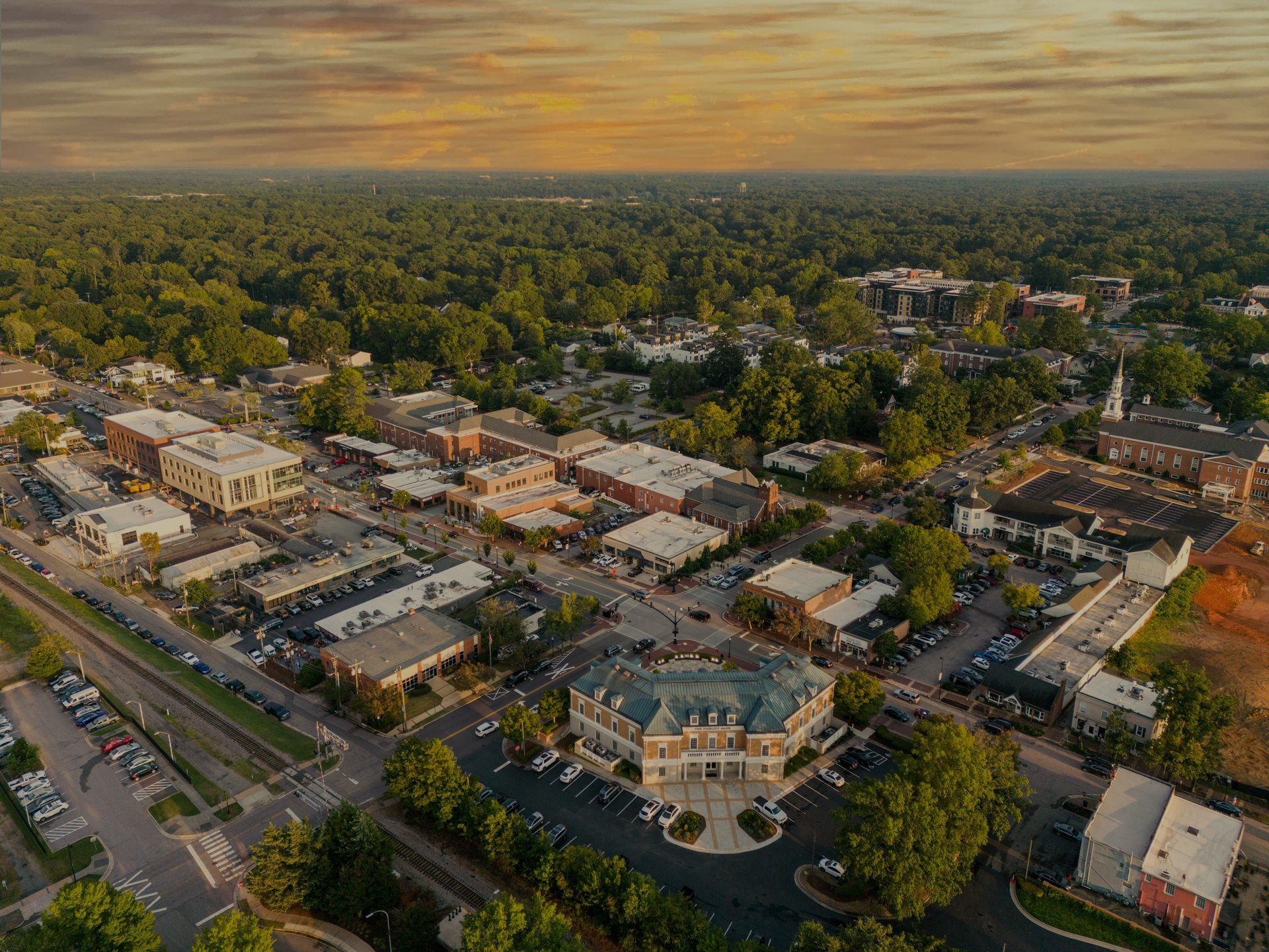 Aerial view of a town with buildings, roads, and surrounding trees under a sunset sky.