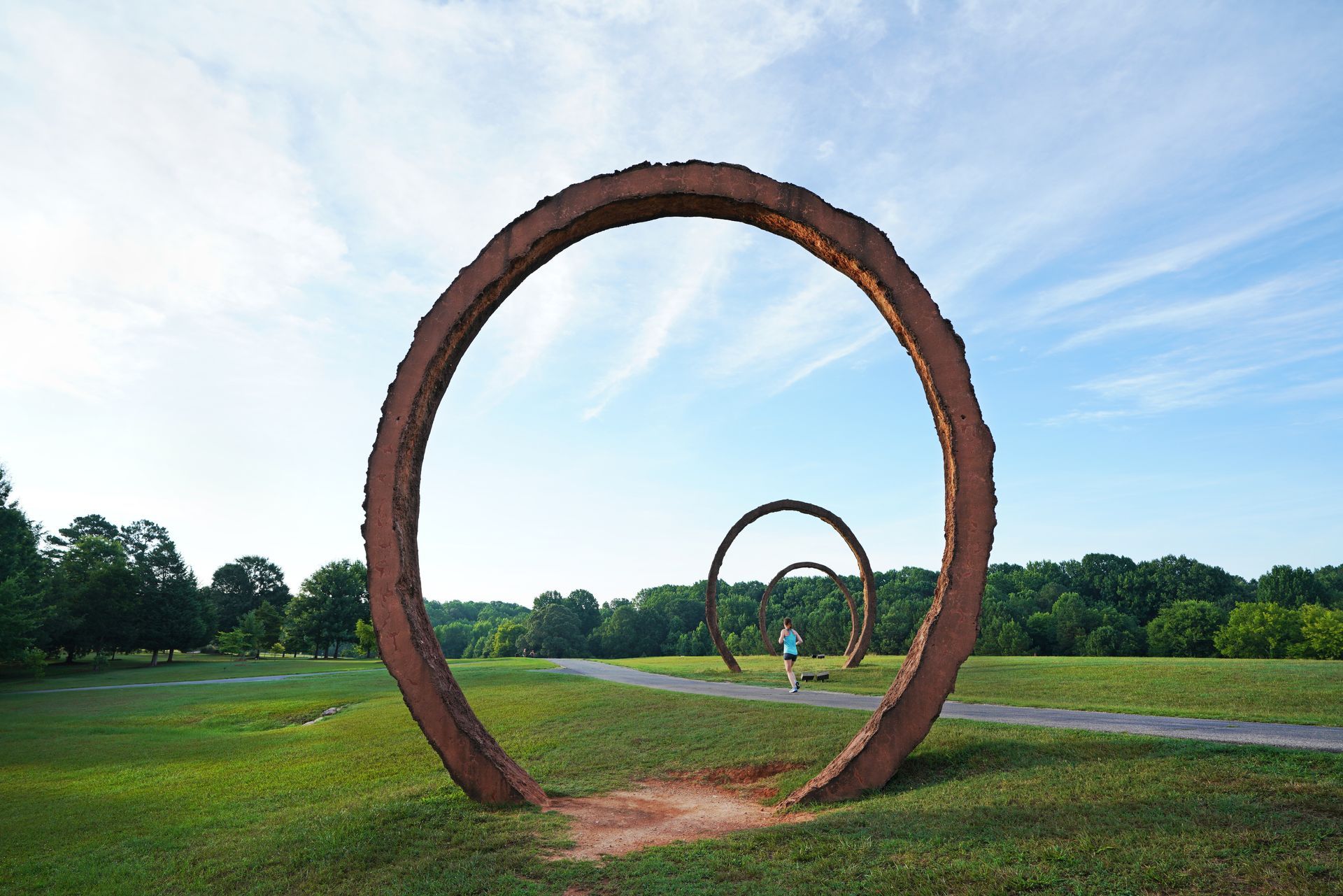 Sculptural steel rings frame a grassy path in a park; a person runs toward the distance under a blue sky.