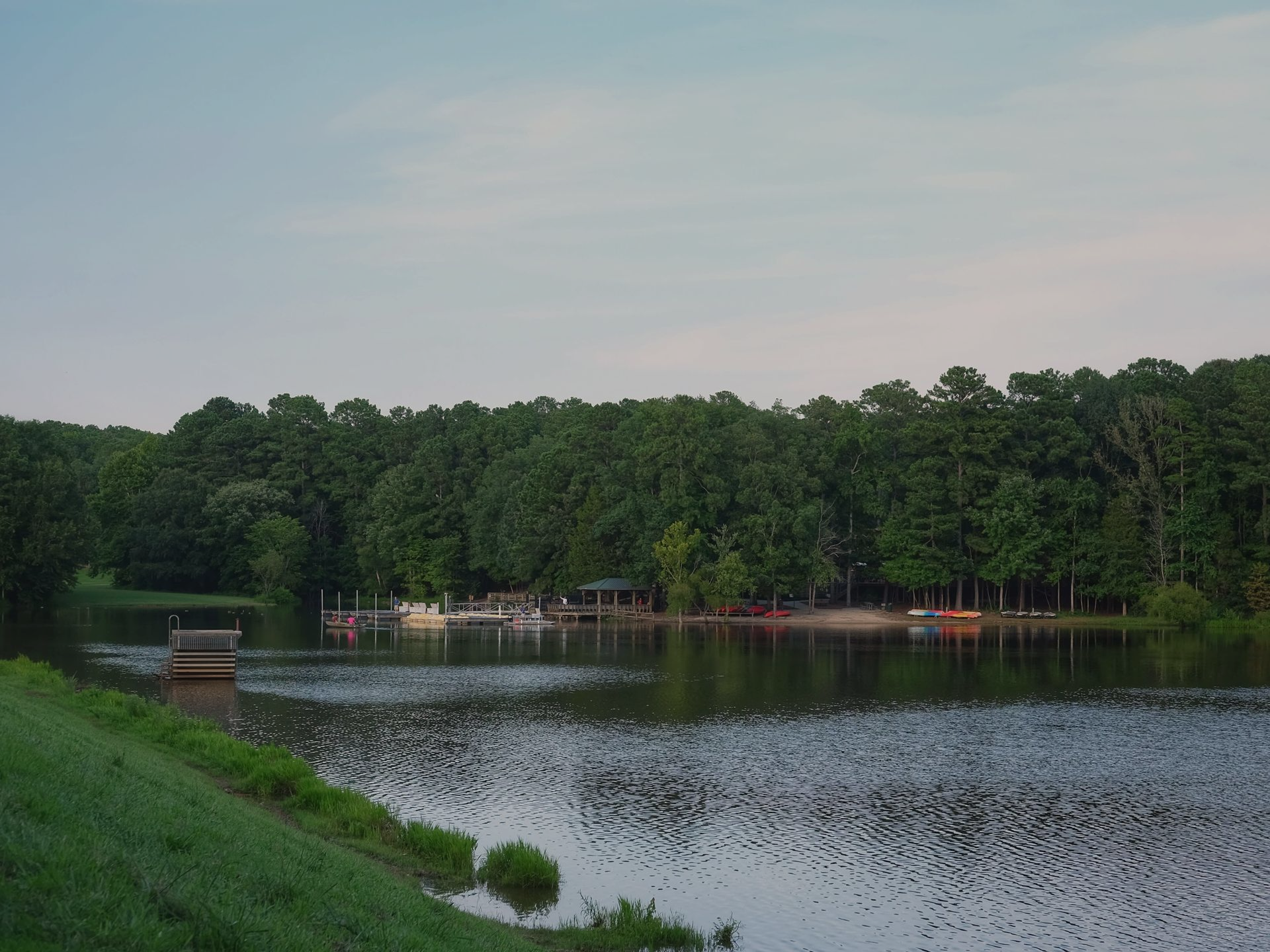 Lake surrounded by green trees, with a dock and small boats visible. Overcast sky.