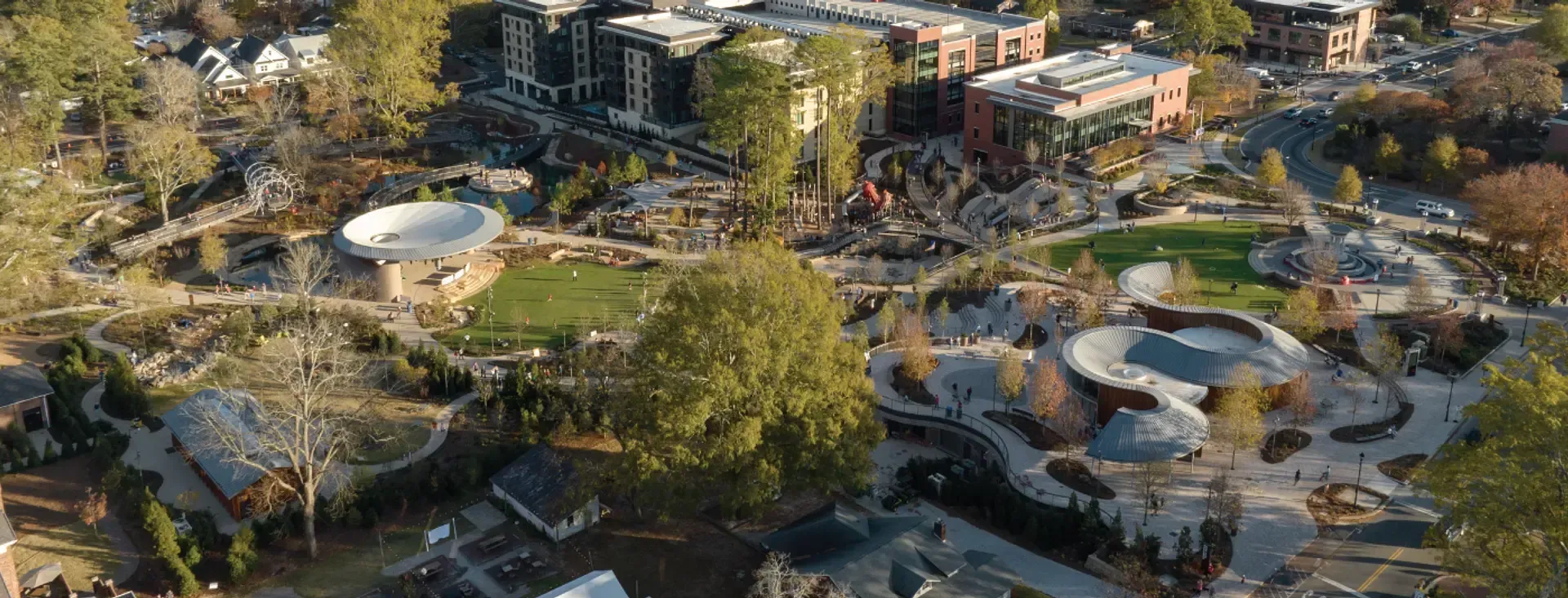 Aerial view of a park with stone paths, greenery, buildings, and a large round structure.