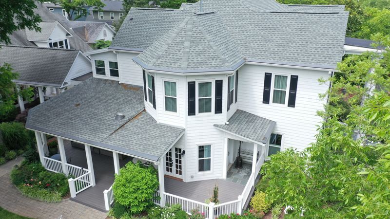 White two-story house with gray roof, porch, and shutters, surrounded by green trees and landscaping.
