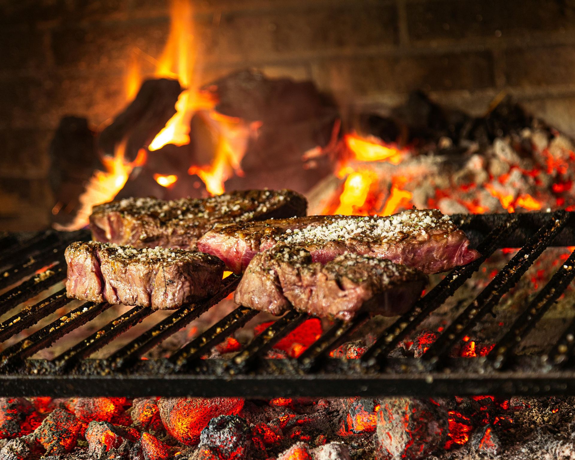 Steaks grilling over hot coals in front of a blazing fire.