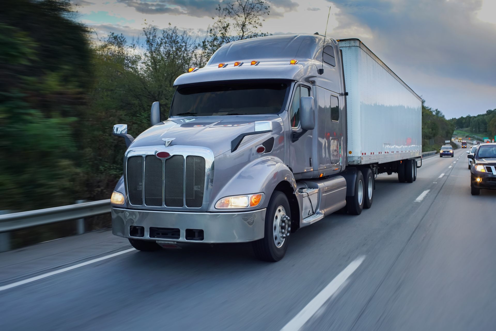 Silver semi-truck driving on a highway beside a guardrail, with another vehicle behind it.