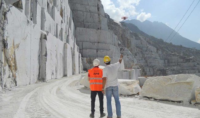 Two men are standing in a quarry looking at a drone flying overhead.