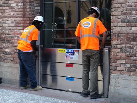 Two men wearing orange safety vests are working on a flood barrier