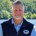 A man with a beard is wearing a black vest and a blue shirt.