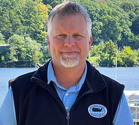 A man with a beard is standing in front of a body of water.