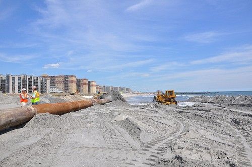 Two men are standing on a sandy beach next to a large pipe.