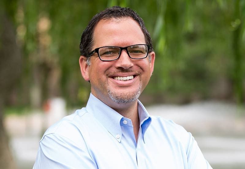 Man in light blue shirt and glasses smiling outdoors, near trees.