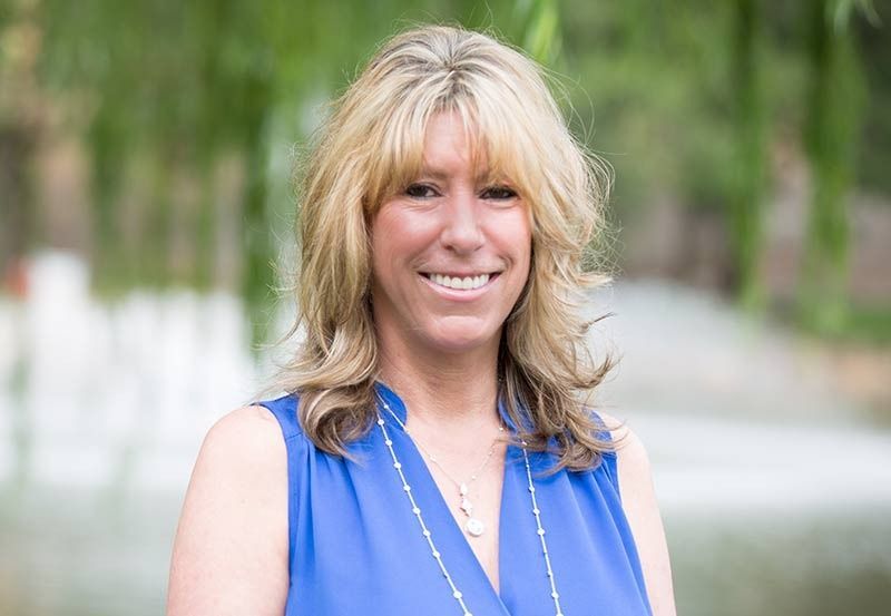 Woman with blonde hair, smiling, wearing a blue top, posing outdoors near a pond and greenery.