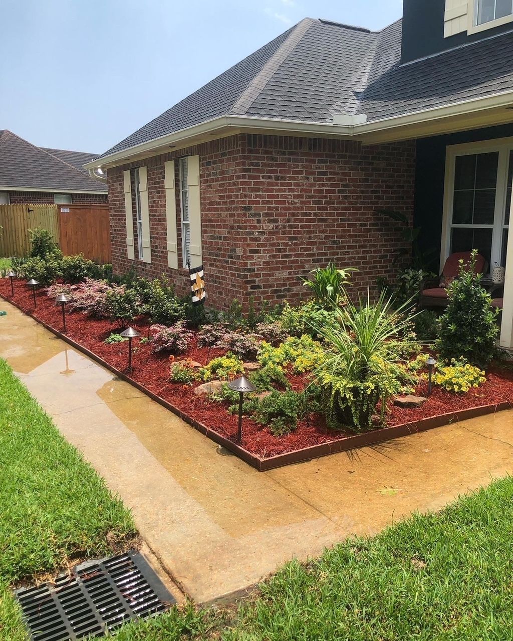 A house with a red brick exterior and a landscaped bed with red mulch, various plants, and pathway lights.