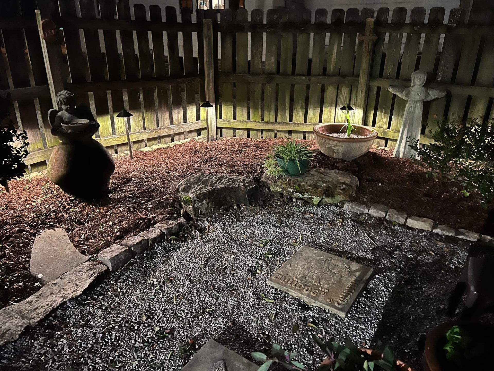 A nighttime garden scene with a stone angel statue, brick-lined beds, and solar lights along a wooden fence.