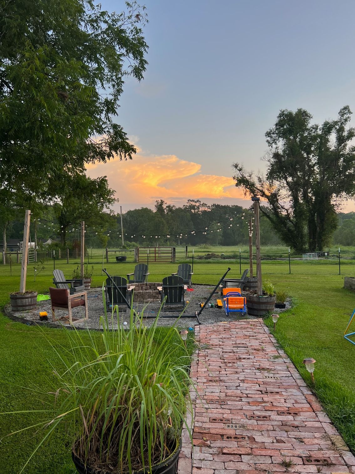 Sunset over a backyard fire pit area with Adirondack chairs. A brick pathway and green lawn surround it.