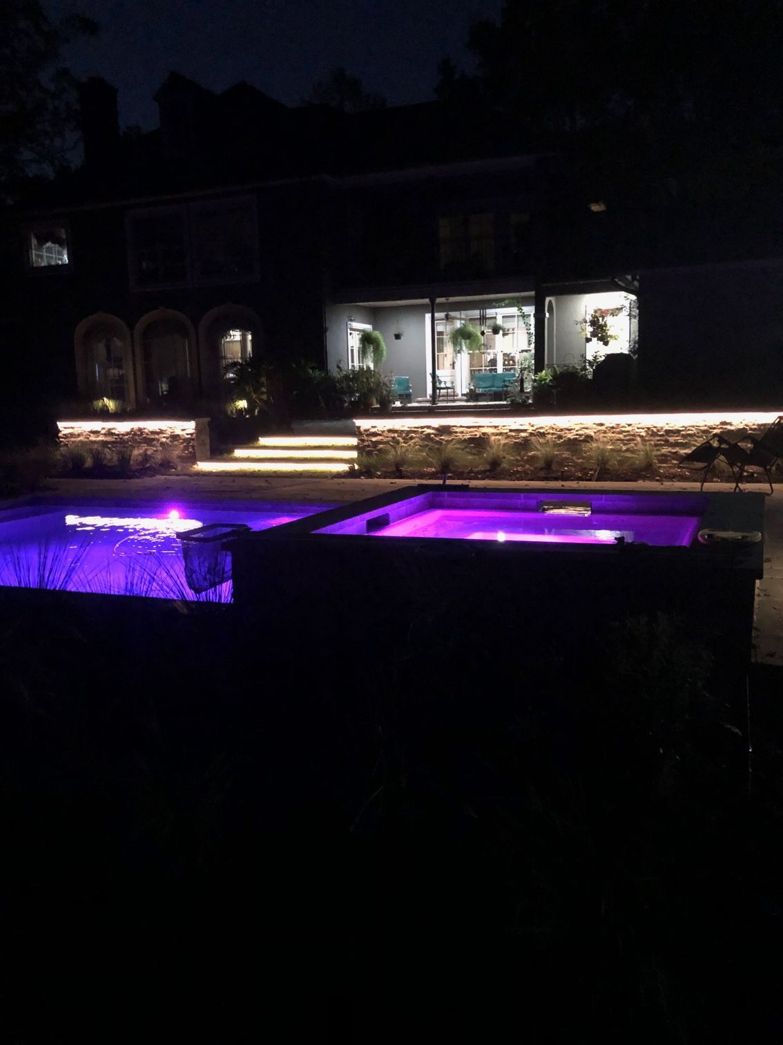 Nighttime view of a luxury pool and spa with purple underwater lights, a house in the background, and subtle lighting on a stone wall.