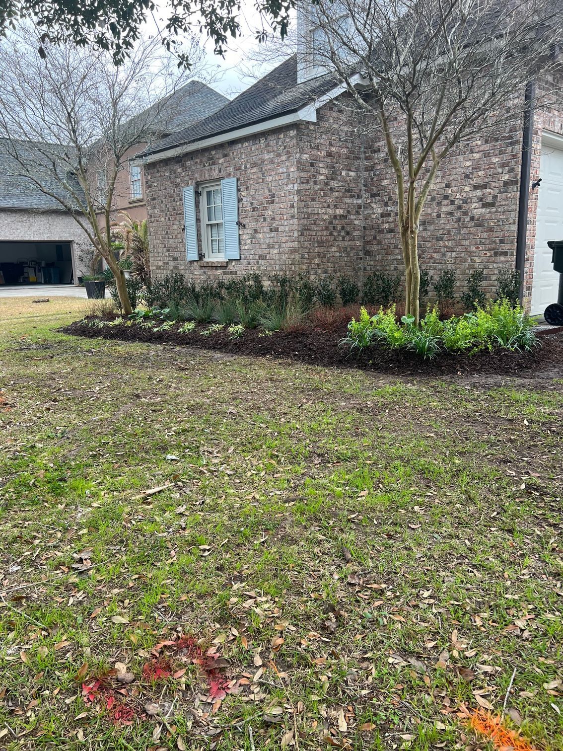 A brick house with a landscaped front yard. Bare trees and green plants are in front of the house. The lawn is patchy green and brown.