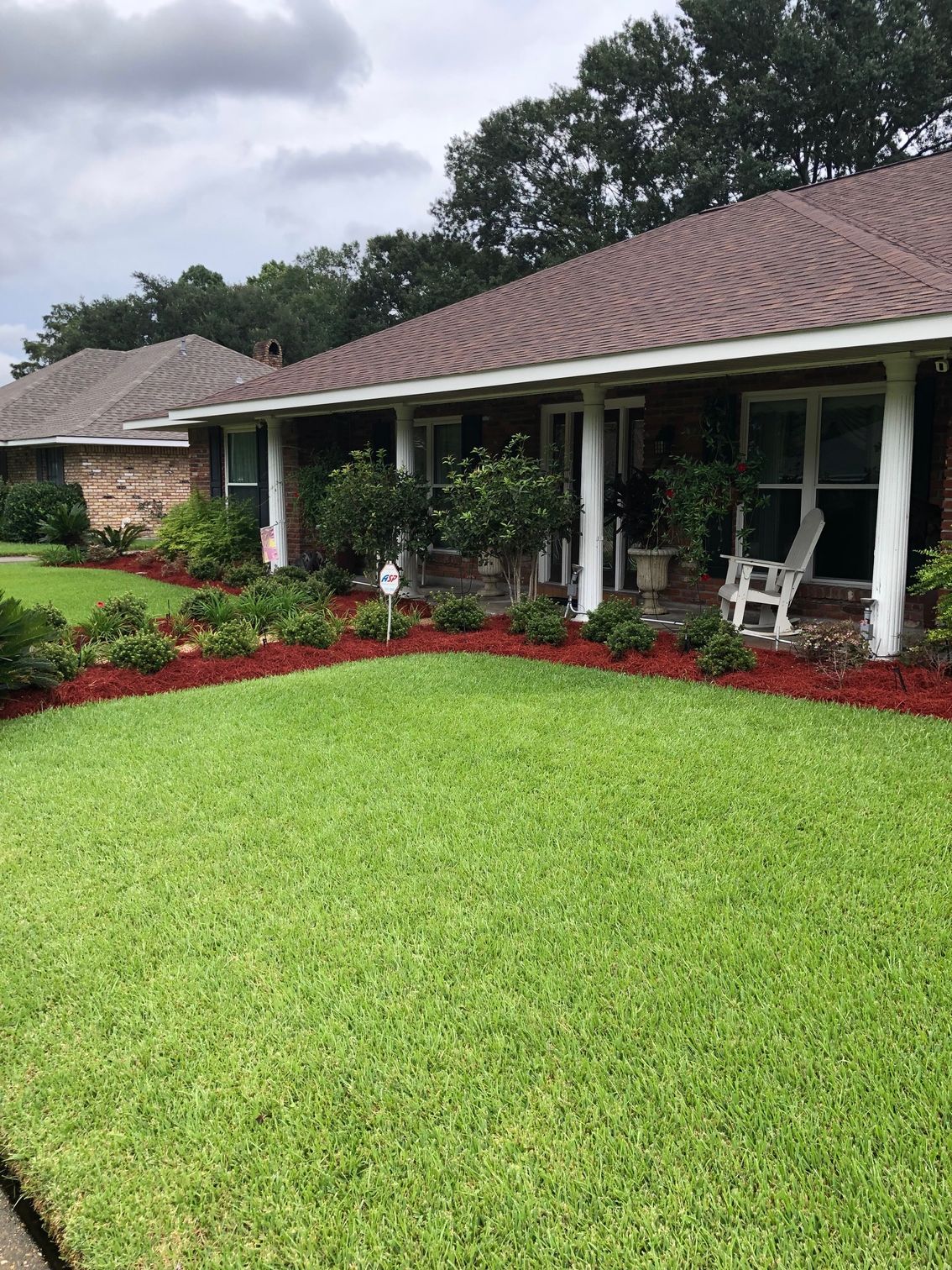 A well-manicured lawn in front of a brick house with a brown roof and red mulch landscaping. White pillars support the front porch.