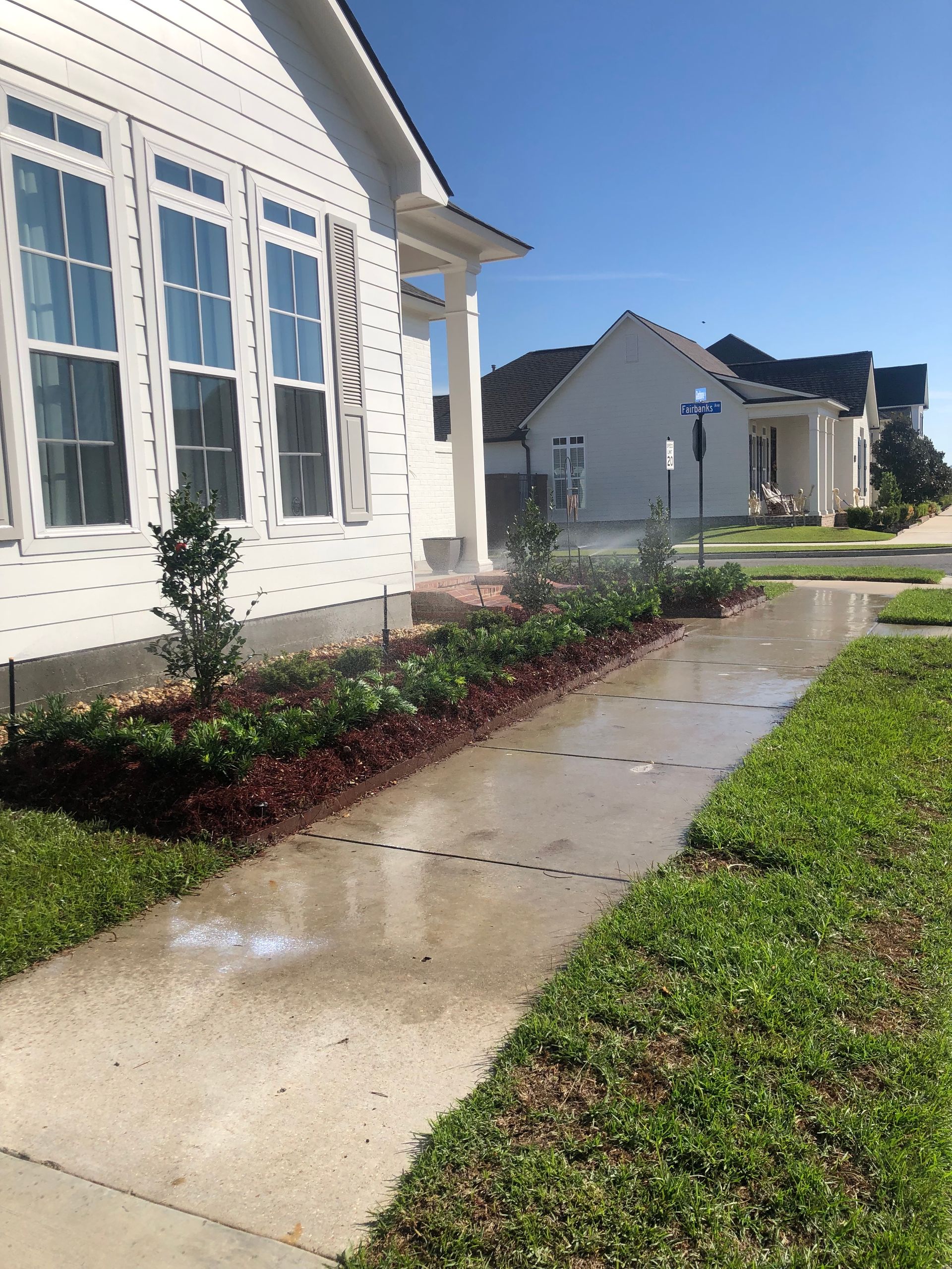 A white house with a sidewalk in front and a flower bed with brown mulch. Several other houses are visible in the background.