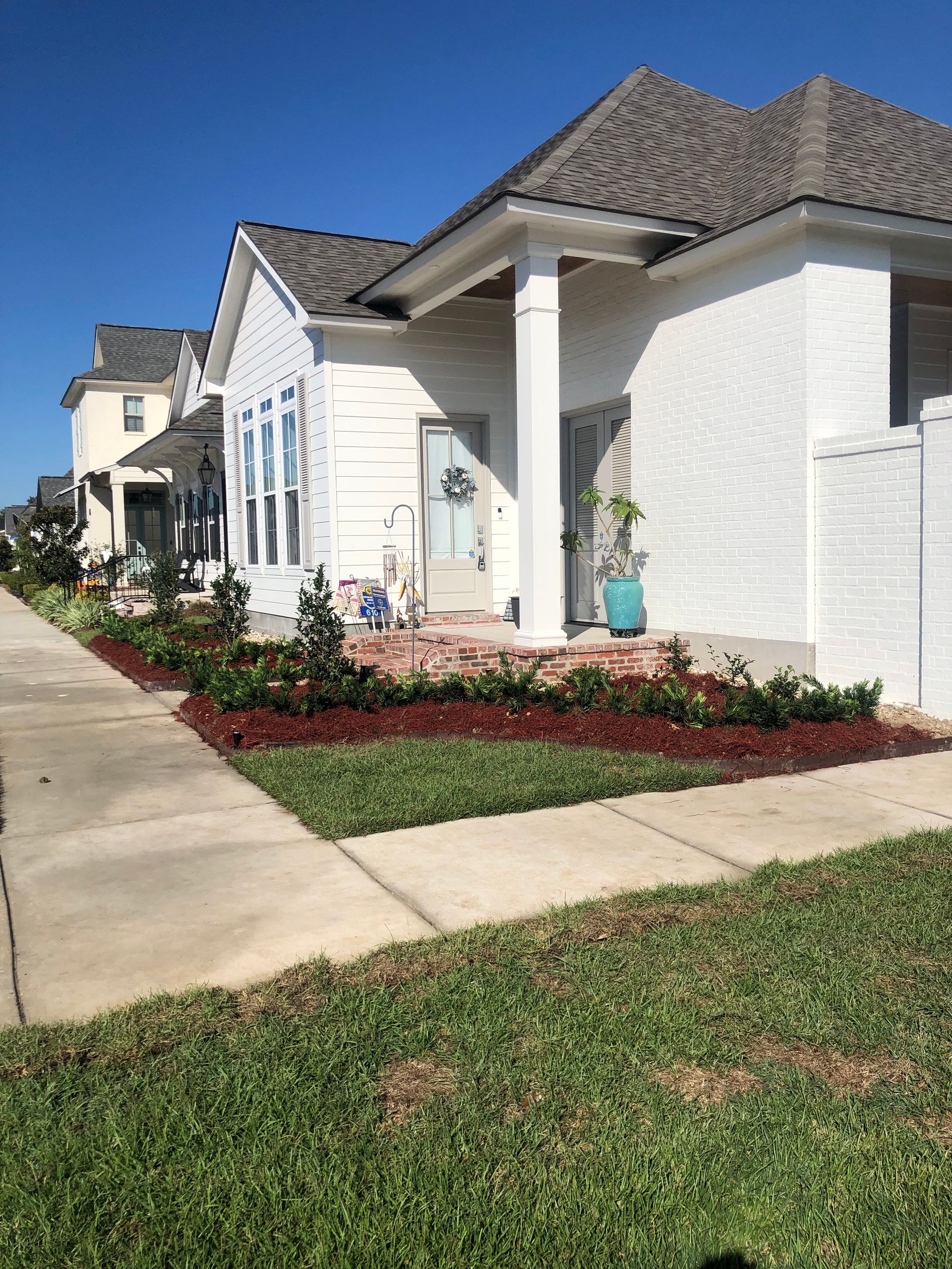 White house with brick accents and a porch, front yard with red mulch and green plants along a sidewalk.