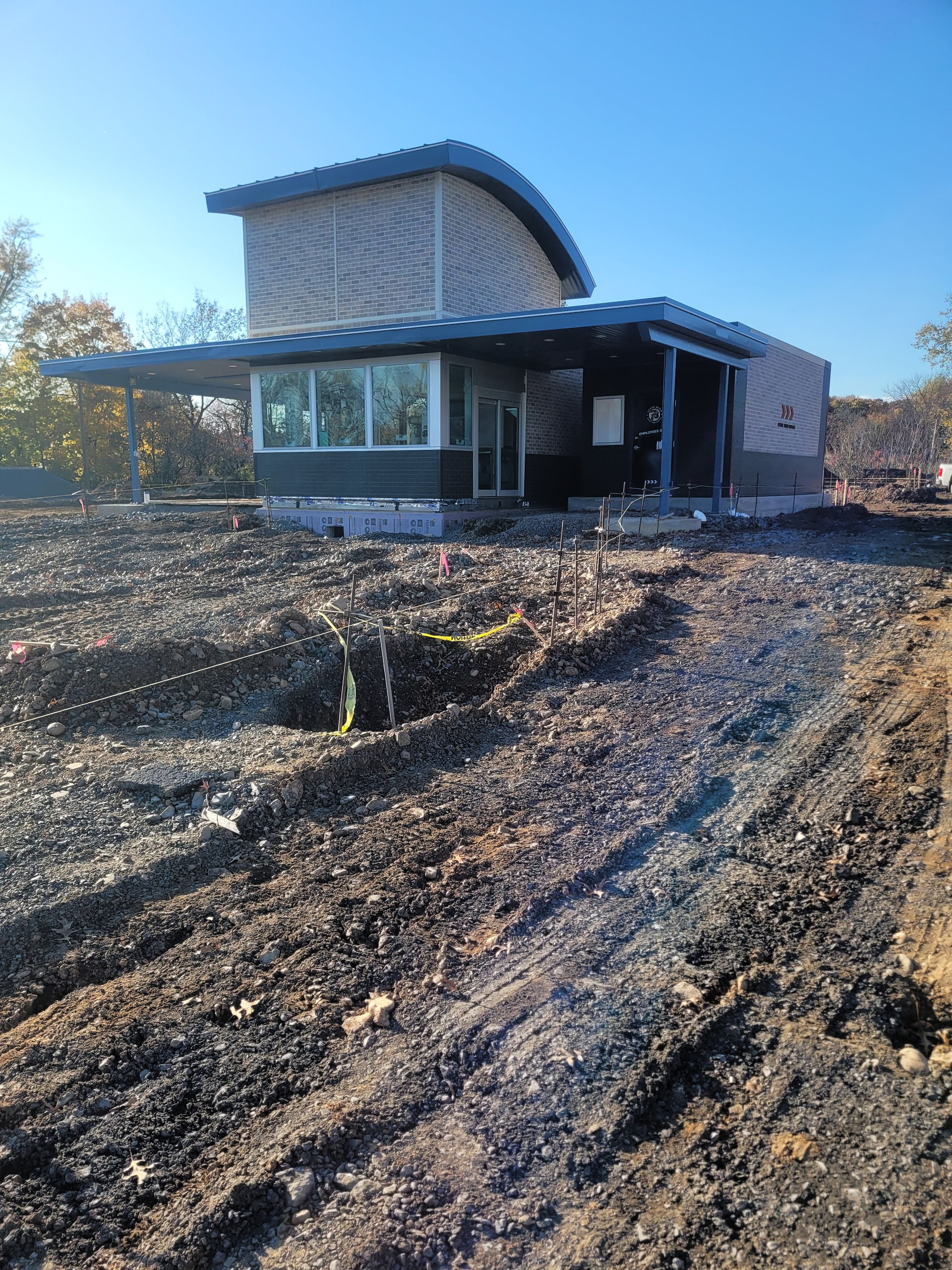Modern building under construction with curved roof, blue accents, set on a dirt lot under a clear blue sky.