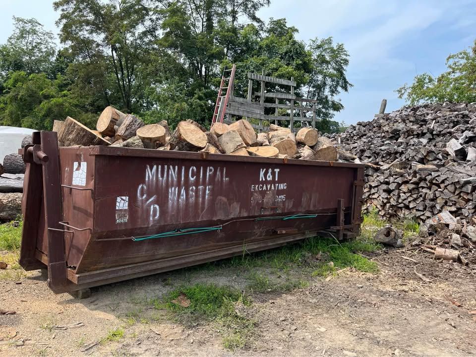 A brown metal dumpster filled with logs. A pile of logs is on the right.
