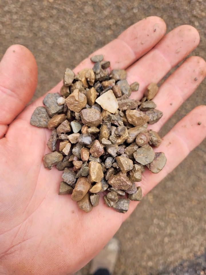 Hand holding a pile of small, irregularly shaped 1B Landscaping, brown and gray gravel.