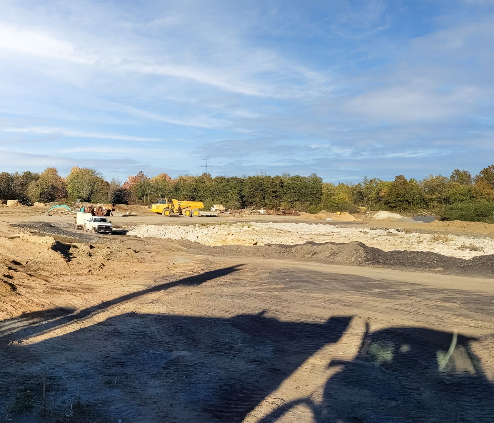 Construction site with earth-moving equipment and a white truck; trees in the background under a cloudy sky.