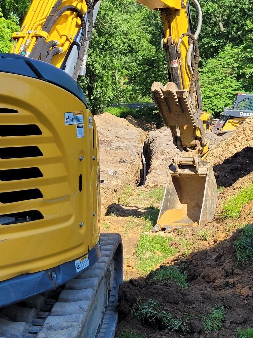 Yellow excavator digging a trench in a grassy area near a wooded background. Construction work is in progress.