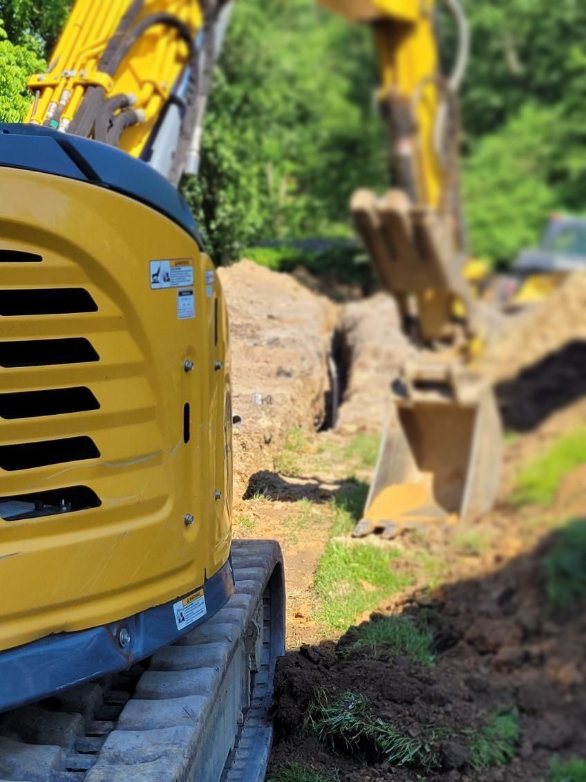 Yellow excavator digging trench in dirt, with another excavator visible in the background.