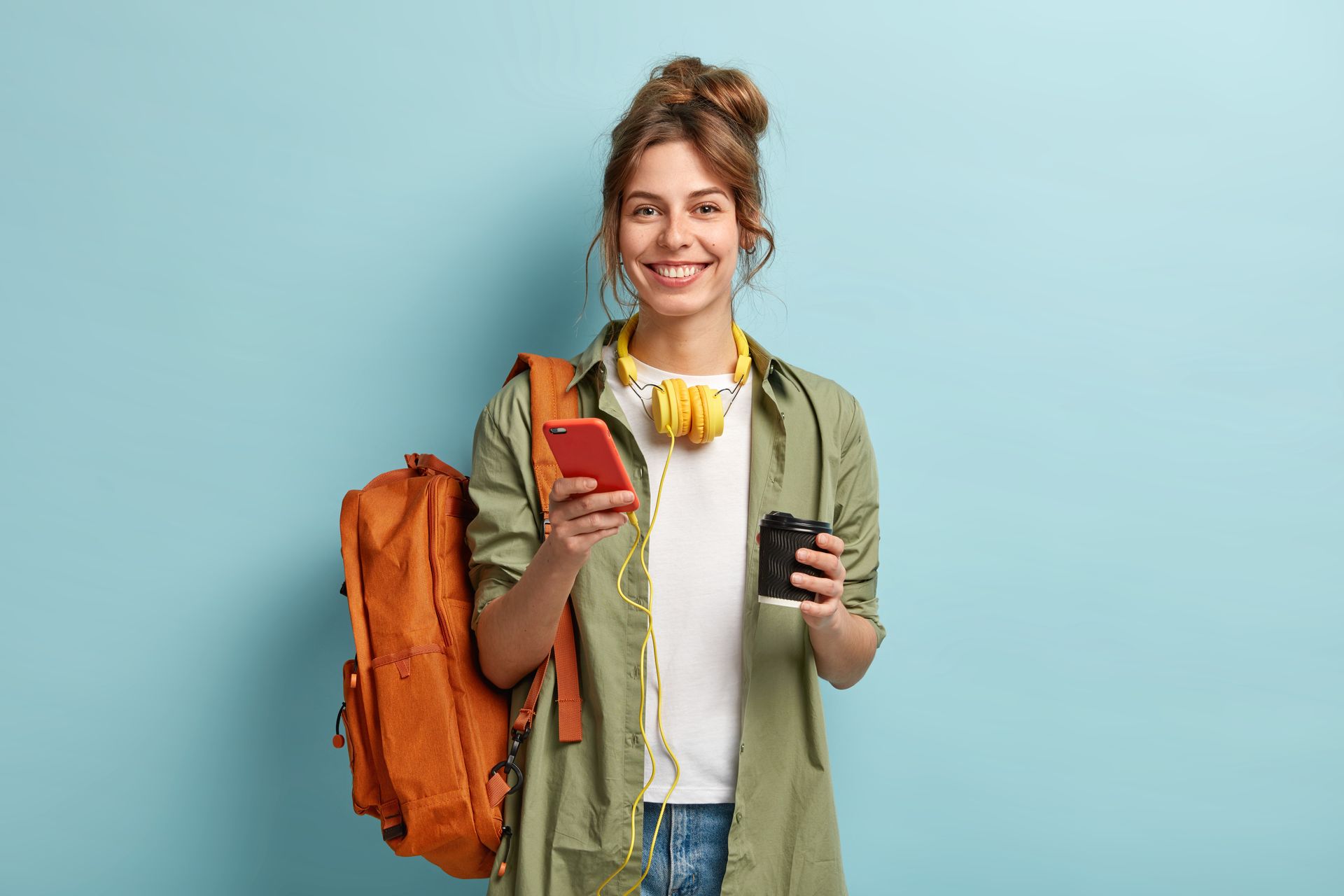 A woman with a backpack is holding a cup of coffee and a cell phone.